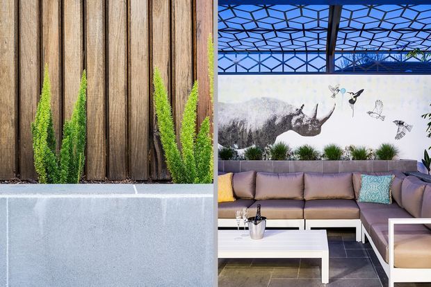 Split image showing a contemporary outdoor lounge area beside a close-up of vertical timber cladding with narrow planted greenery. The lounge features an L-shaped upholstered sectional, white low tables, dark tiled flooring, a botanical and rhinoceros mural, and a geometric steel-and-glass canopy overhead. The overall palette combines warm timber, grey concrete, muted upholstery, and lush green planting.