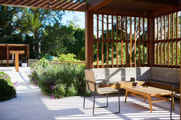 Sunlit outdoor terrace or covered patio with a timber pergola and vertical timber screening, concrete paving, built-in concrete seating, and mid-century-inspired outdoor furniture. The space opens onto a landscaped garden with flowering shrubs and mature trees.