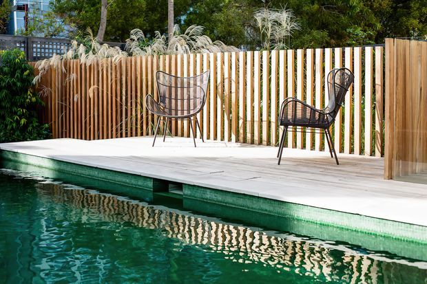 Outdoor swimming pool bordered by a pale timber deck and a vertical timber slat privacy fence, with two black woven lounge chairs and ornamental grasses. The scene has a relaxed, natural contemporary outdoor setting with strong reflections in the pool.