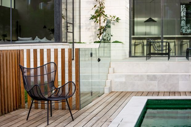 A refined outdoor courtyard and pool area with pale timber decking, a partially visible swimming pool, black wire-frame lounge chairs, a vertical timber privacy screen, and broad pale stone steps leading to a glazed indoor entertaining space. White masonry walls, extensive dark-framed glazing, and minimalist black pendant lights create a contemporary indoor-outdoor setting.