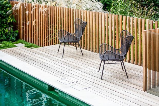 A landscaped outdoor pool deck with pale timber-look decking, two black wire-frame lounge chairs, and a vertical timber slat privacy fence surrounded by lush planting.