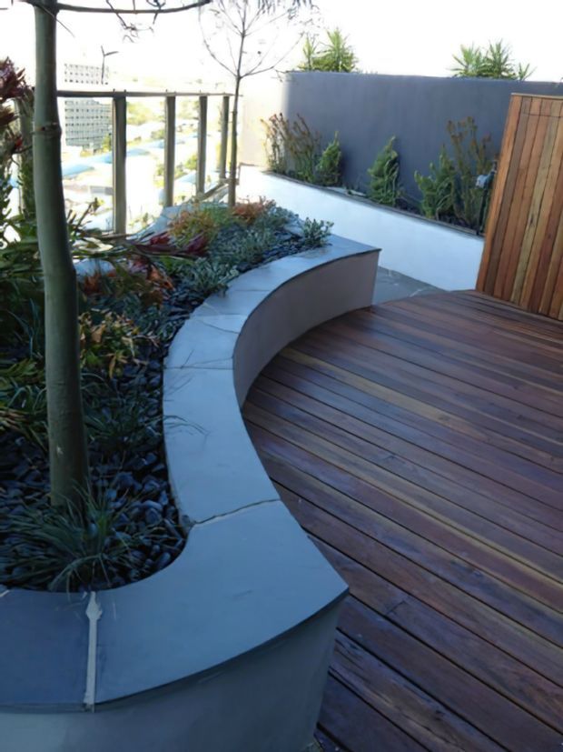 A landscaped outdoor terrace or courtyard with a curved built-in rendered planter and integrated seating edge. The terrace is finished with dark timber decking, slate-like paving around planted beds, and a vertical timber gate or screen. A dark blue-grey boundary wall and glazed balustrade frame the planted space.