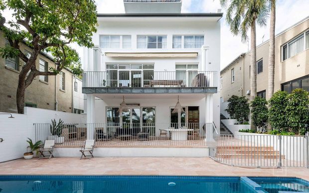 Front exterior view of a multi-storey white rendered residence with balconies, large glazed doors and windows, metal balustrades, landscaped courtyard, tiled pool terrace, and a swimming pool in the foreground. The building has a clean contemporary coastal character and a flat roofline.
