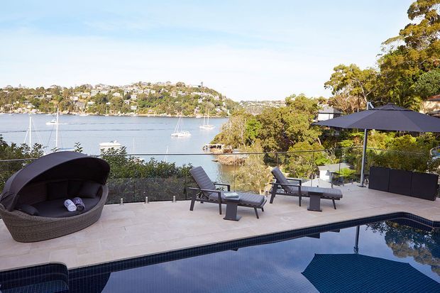 A refined waterfront outdoor entertaining area with a dark-tiled swimming pool in the foreground, pale stone or concrete paving, lounge chairs, a sculptural woven outdoor daybed, large umbrellas, and built-in dark planters. The terrace overlooks a calm bay with boats and tree-covered residential hills beyond.