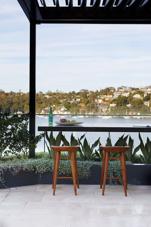 A refined outdoor entertaining terrace with a dark pergola and slim black structural framing, overlooking a leafy waterfront and hillside homes. A narrow stone-topped bar counter with two timber bar stools faces the view, while integrated planter beds provide dense greenery along the edge. Large pale stone pavers create a clean, open floor surface.