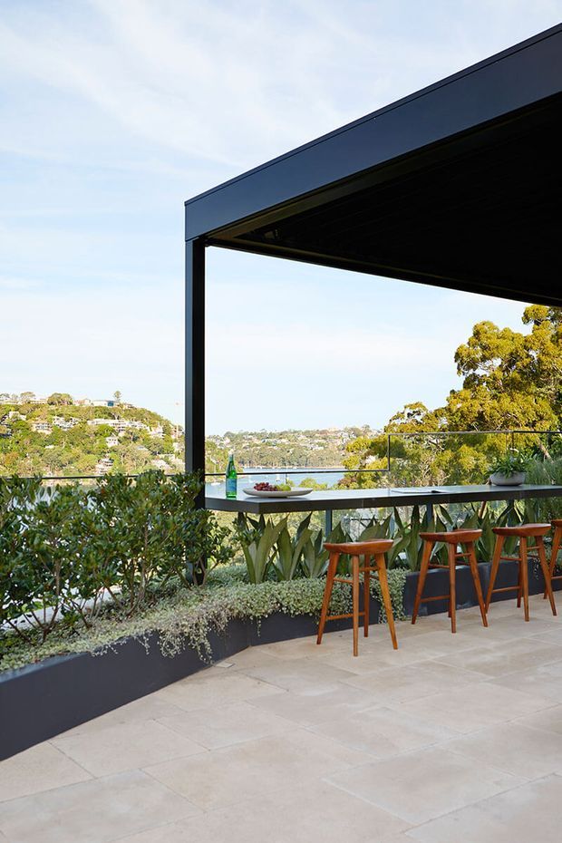 Contemporary rooftop or elevated terrace overlooking a leafy waterfront suburb. The outdoor space features pale large-format paved flooring, dark raised planter beds with layered greenery, a black steel-framed canopy, and a long built-in outdoor bar or counter with timber stools. A glass balustrade and expansive landscape view create an open, refined entertaining area.