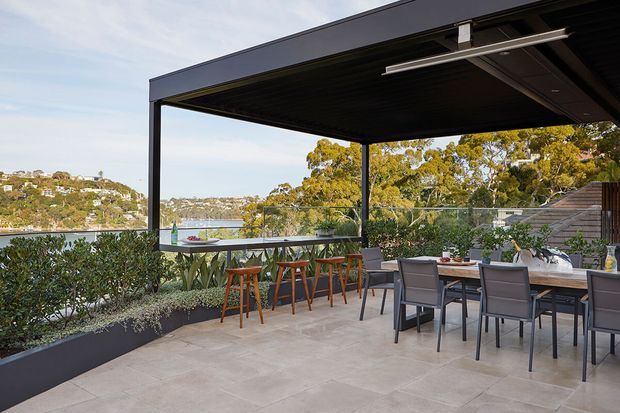 A spacious elevated outdoor terrace overlooking a leafy waterfront setting, with a large black-framed covered pergola, outdoor dining table and chairs, a bar-height counter with stools, built-in planter boxes, and pale tiled paving. The terrace has a contemporary, relaxed entertaining character with expansive open views.