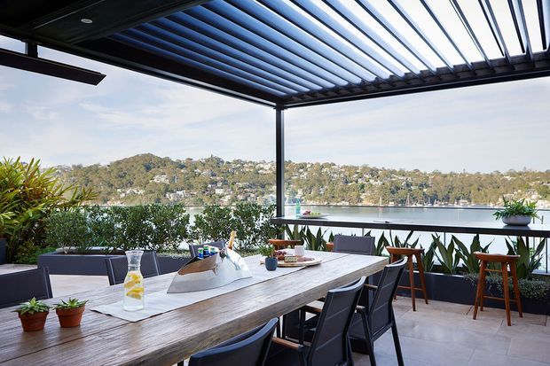 A covered outdoor dining terrace with a large timber dining table, contemporary dark dining chairs, bar stools, planter boxes and a louvred pergola roof. The terrace overlooks a calm body of water and a densely vegetated residential hillside. The space is bright, open and suited to entertaining.
