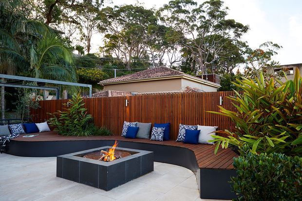A landscaped outdoor entertaining area with a curved built-in bench, timber decking, tiled paving, and a square fire pit. Tropical planting, a timber privacy fence, and a tiled-roof structure create a lush residential courtyard atmosphere.