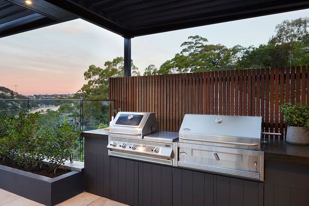A covered outdoor kitchen and barbecue area on a terrace, with stainless-steel grills set into dark cabinetry, a timber slat privacy screen, glass balustrade, planter boxes, and views over a leafy waterfront landscape at dusk.