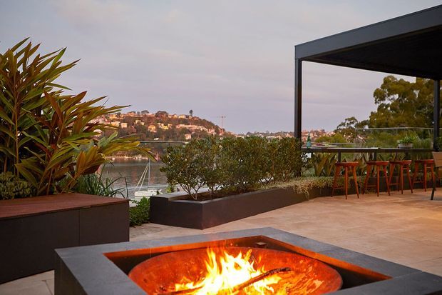 A landscaped waterfront terrace with a built-in rectangular fire pit in the foreground, raised dark planter beds, tropical planting, and a covered outdoor bar or dining pavilion with stools. Calm water and a residential hillside are visible beyond the terrace.
