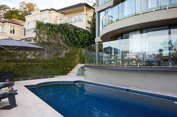 A contemporary multi-level home overlooks a tiled swimming pool and paved entertaining area. The house features extensive curved floor-to-ceiling glazing, glass balustrades, a rounded balcony, and a glazed indoor dining or living area. Mature clipped hedges and ivy-covered walls provide privacy, while a neighbouring traditional residence is visible beyond.