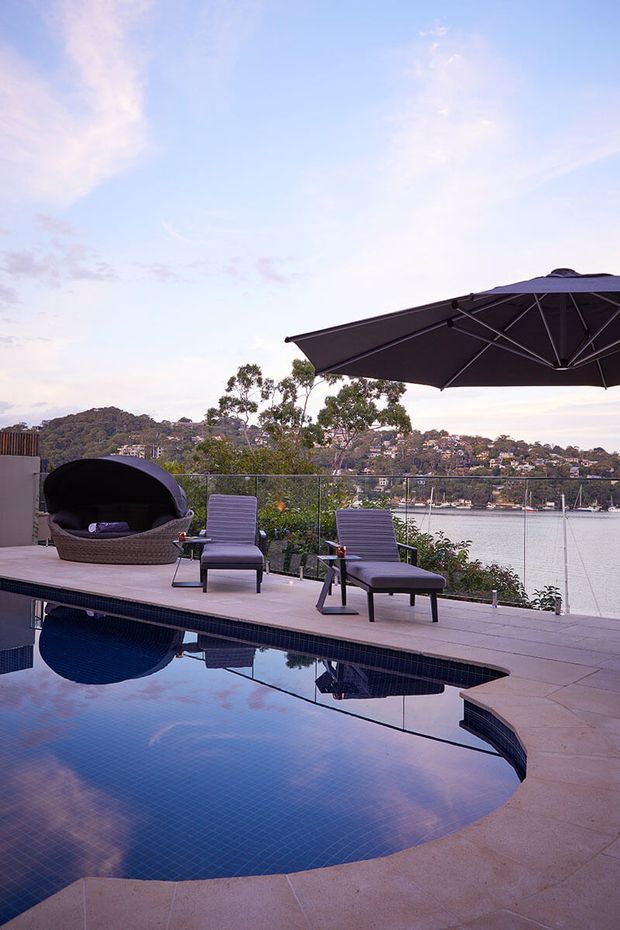 A waterfront outdoor terrace with a curved swimming pool in the foreground, paved pool deck, outdoor lounge chairs, a large dark cantilevered umbrella, and a woven daybed. Glass balustrades frame views across a calm inlet toward wooded and residential hills. The scene has a relaxed, resort-like atmosphere at dusk.