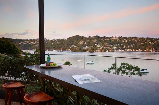 A covered outdoor entertaining area with a long stone-topped bar or bench and timber stools, overlooking a calm waterfront inlet. A glass balustrade, planted edge, small sailboats, and a hillside residential landscape are visible beyond, with soft evening light and pastel sky.