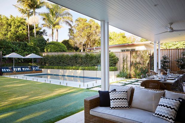 A bright outdoor entertaining area beneath a large covered patio, overlooking a rectangular swimming pool and manicured lawn. The space includes a wicker outdoor sofa with cushions, a dining table and chairs, ceiling fans, striped pool umbrellas, hedges, palms and timber privacy fencing.