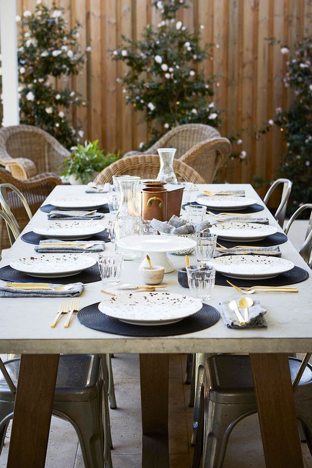 A styled outdoor dining setting with a long rectangular table covered in white tableware, dark woven placemats, cutlery, glasses, candles, and a central copper-toned ice bucket. Woven rattan armchairs and metal dining chairs surround the table, set against a warm vertical timber fence with potted flowering greenery. The overall look is rustic, natural, and inviting.