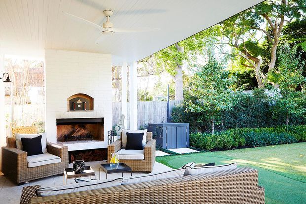 A bright covered outdoor entertaining area with a white-painted ceiling, ceiling fan, substantial white brick fireplace, and built-in wood-burning firebox. Wicker lounge chairs and a large wicker sofa surround a low coffee table, opening onto a manicured green garden with hedging and mature trees. The space feels like a relaxed, refined outdoor living room.