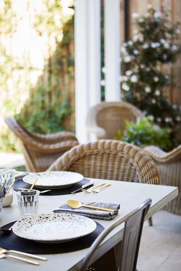 Bright, softly focused dining setting with a pale rectangular table dressed with speckled ceramic plates, dark placemats, clear glasses, cutlery, and folded grey napkins. Woven rattan dining chairs surround the table, with large white-framed glazed openings and leafy greenery creating an airy indoor-outdoor atmosphere.