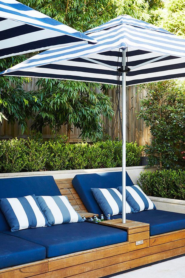 A landscaped outdoor seating area with built-in navy upholstered loungers and striped cushions beneath large navy-and-white striped umbrellas. The seating is integrated into a warm timber deck and surrounded by clipped hedges, trees, and a timber privacy fence, creating a resort-like garden setting.