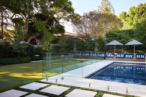A landscaped outdoor pool area with a rectangular swimming pool, clear frameless glass safety fencing, pale paved surrounds, stepping-stone paths, manicured lawn, dense hedging and mature trees. Blue-and-white striped sun loungers and parasols line the far side of the pool, creating a polished resort-like setting.