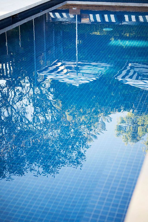 Close-up view of a clear blue swimming pool with a blue tiled interior and pale coping along the edge. Reflections of surrounding trees, sky, and a nearby structure ripple across the water surface.