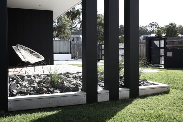 Contemporary residential courtyard viewed from a covered outdoor area, featuring bold black vertical posts, dark wall panels, raised timber-edged rock garden beds with ornamental grasses, clipped lawn, and a white wire lounge chair. A fenced garden and neighboring suburban structures are visible beyond.