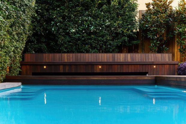 A landscaped residential pool area with a bright blue swimming pool in the foreground, bordered by dark timber-clad retaining walls and built-in bench seating. Small integrated lights illuminate the timber wall, while dense greenery and established planting surround the space, creating a private outdoor setting.
