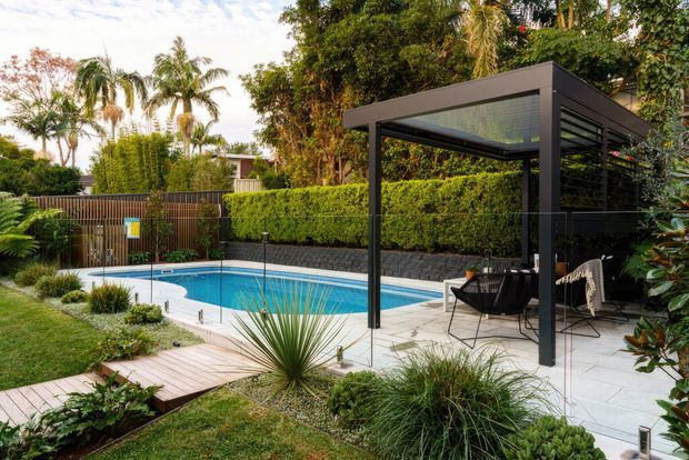 A landscaped residential backyard featuring a rectangular swimming pool, pale paved pool terrace, timber garden path, clipped hedges, tropical planting, and a contemporary black metal pergola with a translucent roof and outdoor seating.