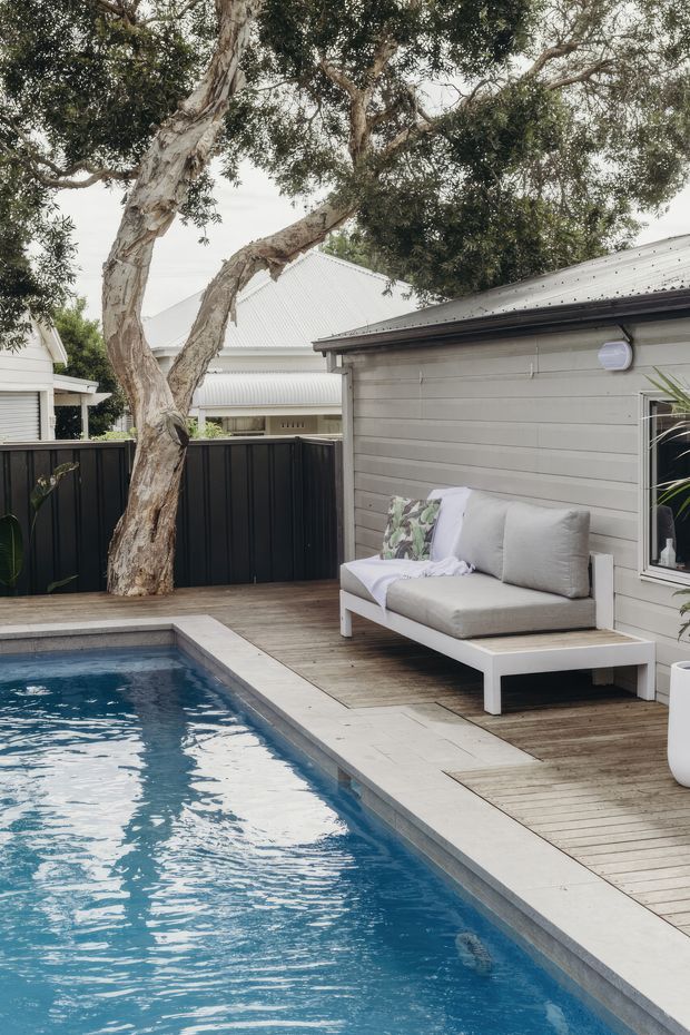 A residential outdoor pool area with a narrow rectangular swimming pool, pale stone coping, and timber decking. A white outdoor sofa with grey cushions sits beside a light grey weatherboard-clad building, enclosed by a dark vertical fence. Mature tree canopy provides shade, creating a relaxed backyard setting.