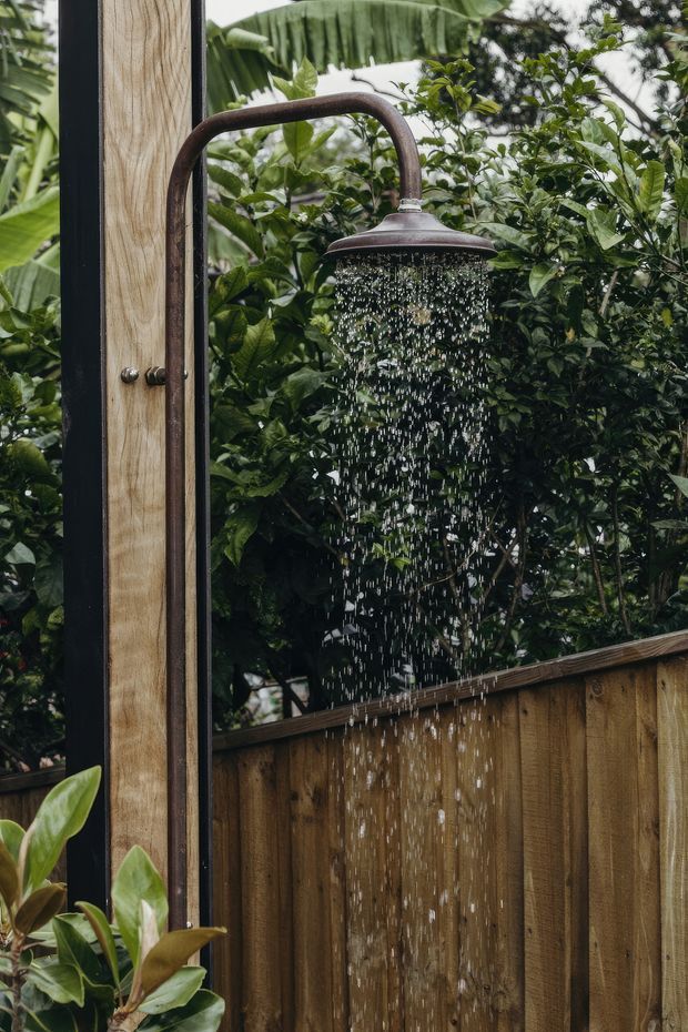 Close-up photograph of an outdoor shower with a dark aged-metal rainfall showerhead and pipe mounted beside a vertical timber privacy screen. Water is visibly cascading from the showerhead, with dense tropical foliage forming the background and a timber fence below.