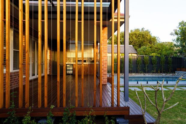 Exterior view of a contemporary brick home with a timber deck and covered outdoor area screened by closely spaced vertical timber slats. The deck overlooks a backyard swimming pool, lawn and fencing, with large glazed doors and warm timber detailing visible along the facade.
