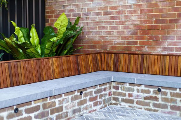 Close-up of a built-in outdoor corner bench against a red brick wall. The bench has warm vertical timber slat cladding and a pale grey concrete or stone seat, with brickwork continuing below and grey brick paving underfoot. Tropical foliage is planted behind the bench, and a dark-framed opening is partially visible at left.