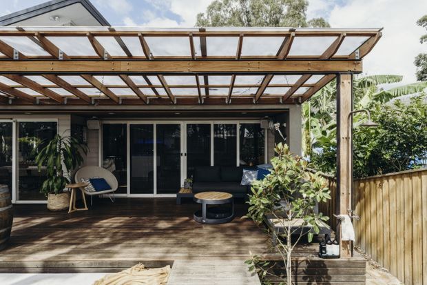 A covered timber deck adjoining a contemporary home, with a transparent pergola roof, timber decking, glass sliding doors, outdoor lounge seating, potted plants and a timber privacy fence. The space is bright and leafy, creating a relaxed outdoor living area.