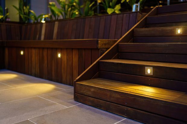 A warmly lit exterior or semi-outdoor stairway with broad dark timber steps and matching vertical timber planter cladding. Small integrated step and wall lights illuminate the stairs, while tiled paving and lush planting frame the scene.