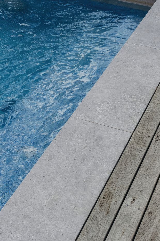 Close-up view of a swimming pool edge with vivid blue water, pale grey concrete or stone coping, and weathered timber decking along the side. The image focuses on the pool surround materials and clean geometric junction between paving and deck.
