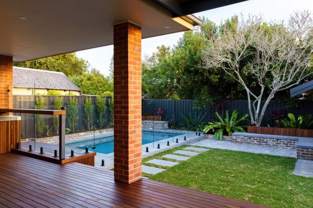 A covered outdoor deck and entertaining area overlooking a compact swimming pool and landscaped courtyard garden. The space features dark timber decking, brick columns, clear glass balustrades, a dark metal boundary fence, raised masonry garden beds, stepping-stone paving, and abundant greenery. A neighbouring tiled-roof structure is visible beyond the fence.