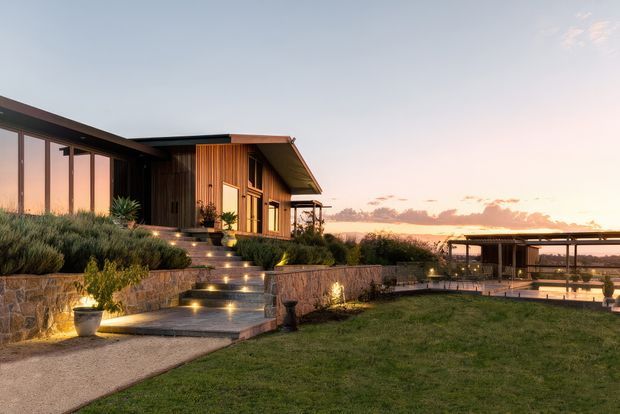 A contemporary single-storey house set in a landscaped elevated site at sunset. The home features extensive floor-to-ceiling glazing, warm vertical timber cladding, dark slender rooflines, broad eaves, and layered stone retaining walls. Illuminated steps lead through native-style planting toward the entrance, while a separate covered outdoor structure and terrace are visible to the right.