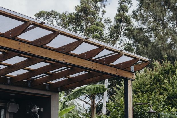 Close-up exterior view of a timber pergola or covered outdoor structure, with exposed dark-stained timber rafters and beams supporting translucent roof panels. Black steel connectors and posts are visible, with lush garden vegetation surrounding the structure.