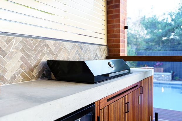 Close-up of a built-in outdoor kitchen beside a swimming pool, featuring a stainless-steel barbecue grill on a light concrete benchtop, timber slatted cabinetry, a tiled backsplash, brickwork, and a timber-framed opening overlooking the pool.