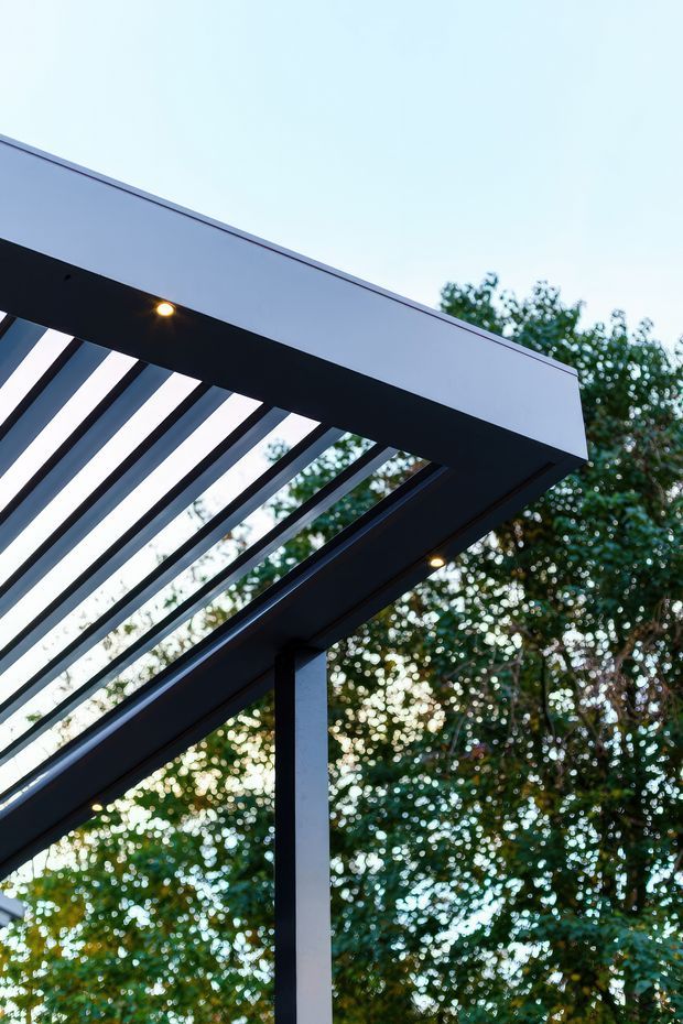 Close-up architectural view of a contemporary outdoor pergola or covered terrace structure, featuring a flat white fascia, dark structural beams, evenly spaced roof slats, integrated recessed lighting, and a dark square support post. Dense greenery is visible behind the structure.