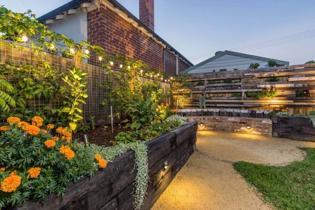 A landscaped residential courtyard or backyard with raised garden beds built from dark timber sleepers, dense planting, warm integrated garden lighting, and a textured timber privacy screen. A red-brick house with a pitched roof and chimney forms the backdrop, with a compact gravel or compacted-earth pathway leading through the garden.
