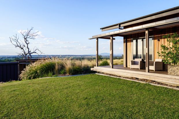 Exterior view of a contemporary single-storey home with extensive timber cladding and a covered outdoor terrace. Full-height glazed sliding doors open onto a timber deck with outdoor seating, overlooking a landscaped lawn and open rural landscape. The building features a low-pitched roof, strong horizontal lines, dark structural framing, and a warm natural-material palette.