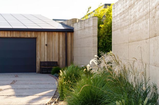 Exterior view of a contemporary garage or outbuilding with vertical timber cladding, a dark garage door, a low-pitched metal roof, and tall pale concrete or masonry boundary walls. A paved driveway and ornamental grasses form a restrained, modern landscape composition.