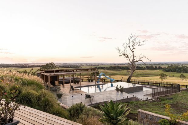 A landscaped rural property viewed from an elevated timber deck, featuring a large paved terrace and swimming pool with a waterslide, shaded outdoor pavilion, dark balustrades, native-style planting, and expansive farmland views beyond. The scene is captured in warm, soft evening light.