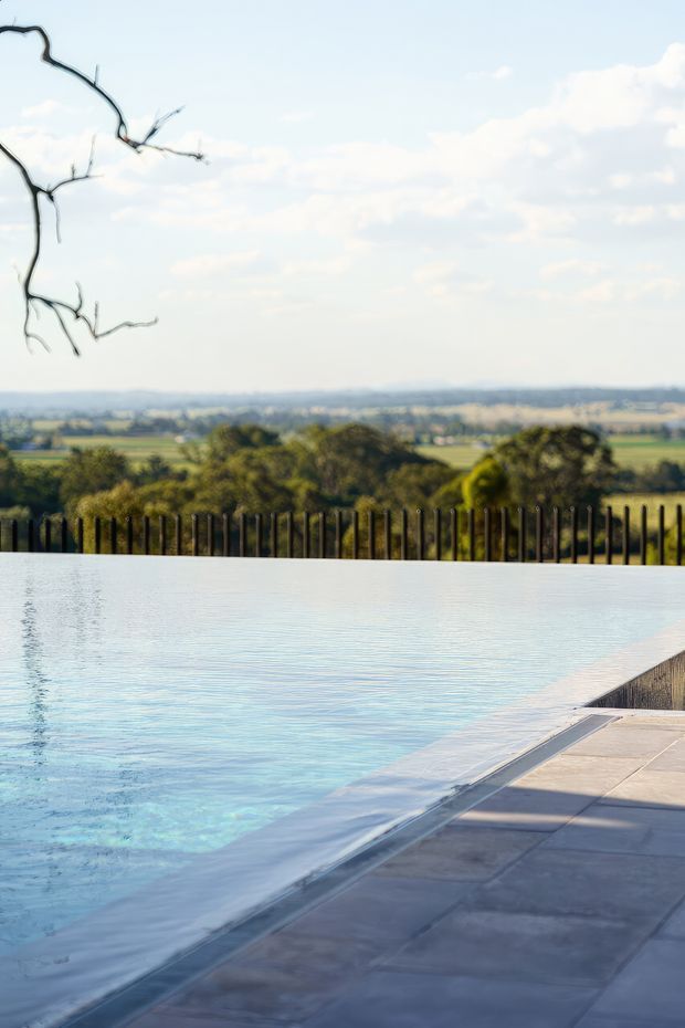 A tranquil outdoor infinity-edge swimming pool occupies the foreground, with pale blue water meeting a dark tiled or stone pool surround. A black vertical metal fence borders the property, beyond which rolling rural farmland and trees extend under a bright, lightly clouded sky. Bare branches frame the upper left of the view.