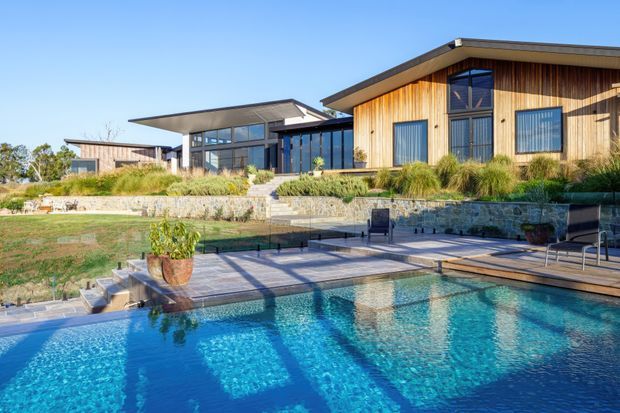 A contemporary single-storey timber-clad residence viewed from the landscaped pool area. The home features broad low-pitched roof planes, extensive dark-framed glazing, a stone retaining wall, and a paved pool terrace with a clear blue swimming pool in the foreground.