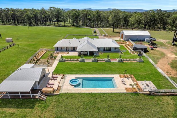 Aerial view of a large single-storey rural property with a light-coloured house, multiple outbuildings, fenced lawns, landscaped garden beds, paved terraces and a rectangular swimming pool with a circular spa. The property is surrounded by open pasture and mature trees, with distant hills visible beyond.