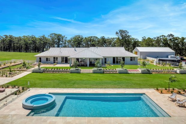 A wide exterior view of a single-storey rural-style residence with a light grey pitched metal roof, pale cladding, covered verandah and landscaped front garden. A large rectangular swimming pool with a circular spa dominates the foreground, with open lawn, mature trees and a separate outbuilding behind the house.
