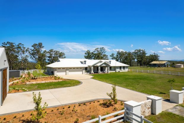 A wide exterior view of a single-storey rural property with a low-pitched light roof, white rendered walls, attached garage, covered entry, broad concrete driveway, landscaped garden beds, fencing and open grassy surroundings under a clear blue sky.