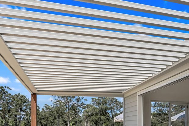 Close-up exterior view of a modern covered outdoor area with a white horizontal slatted pergola roof attached to a light-coloured weatherboard-clad building. Timber posts support the structure, with blue sky and trees visible beyond; part of a glazed opening is visible on the right.